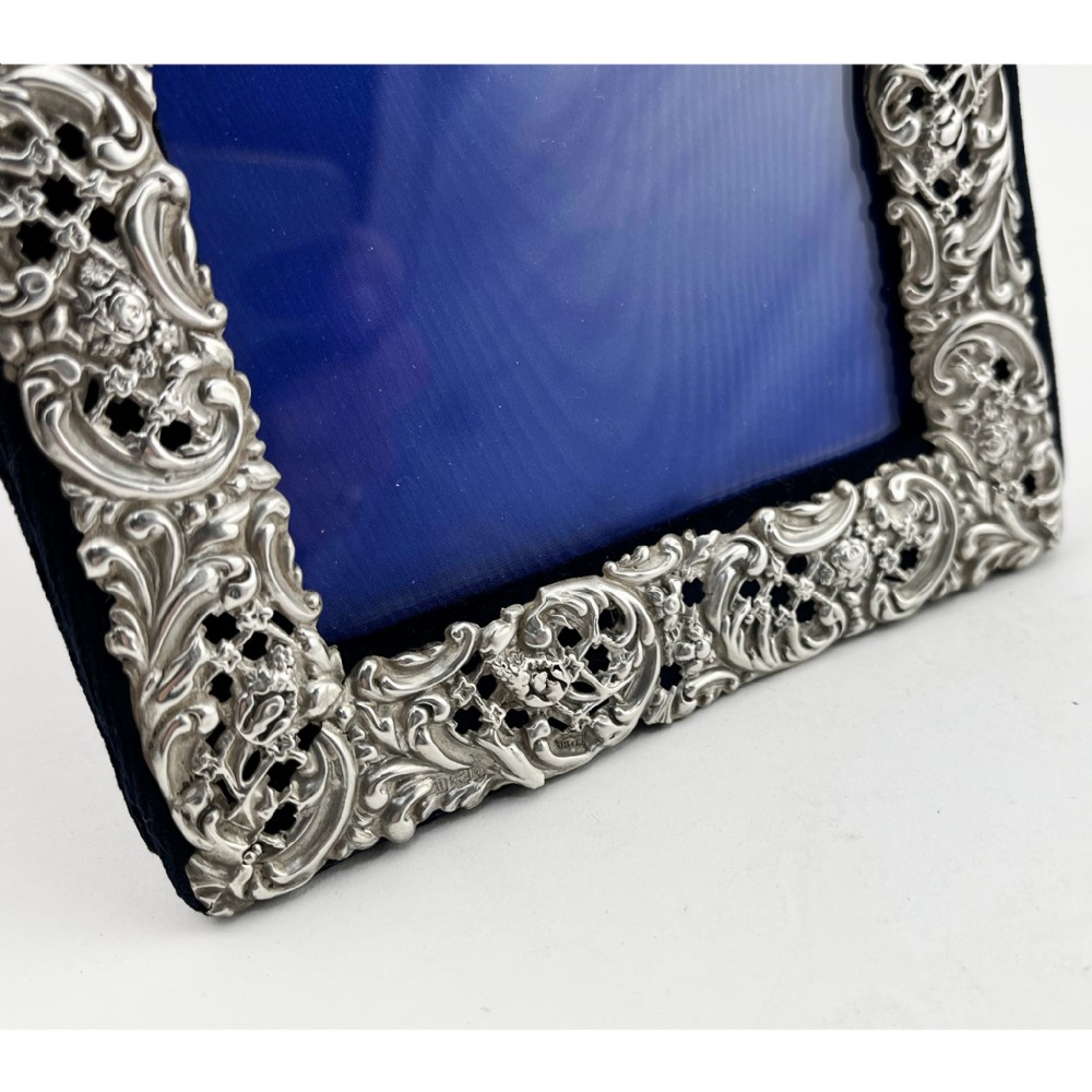 A rectangular sterling silver photo frame featuring intricate floral and scroll repousse metalwork mounted on a dark blue velvet easel backing. View V