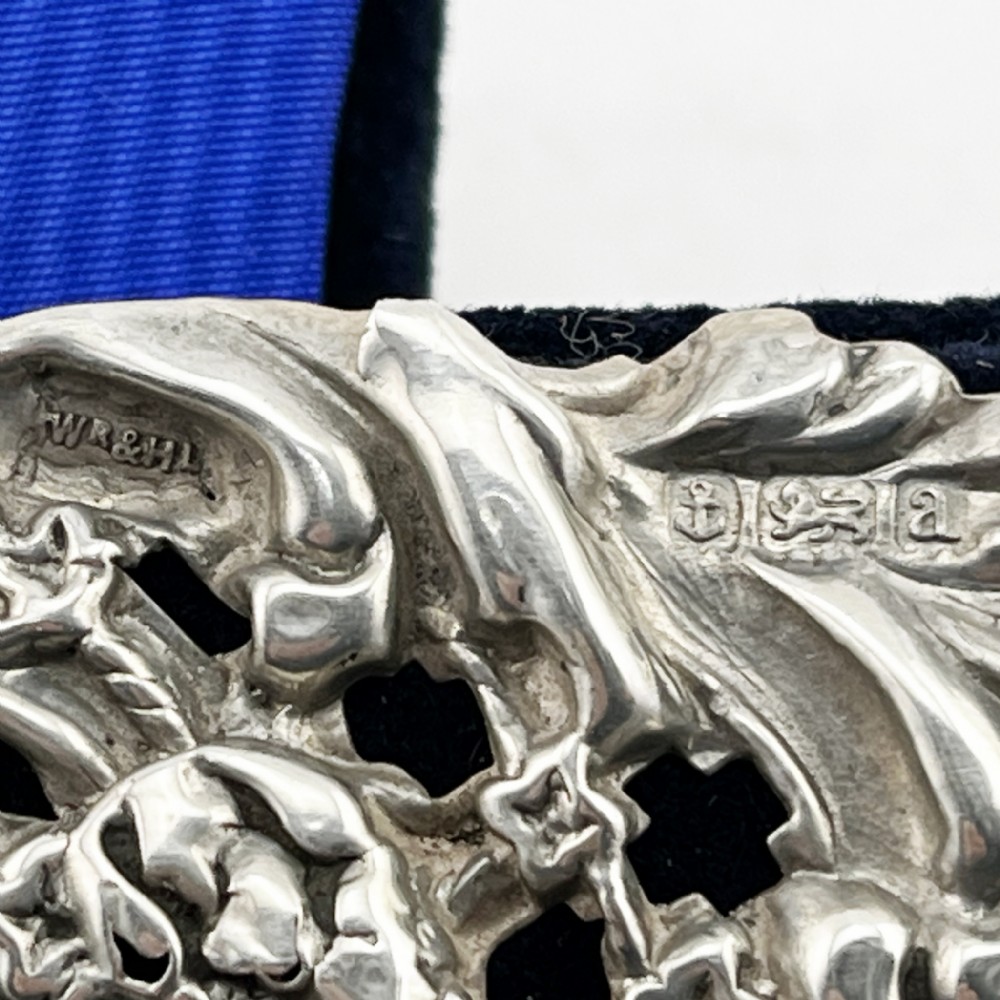 A rectangular sterling silver photo frame featuring intricate floral and scroll repousse metalwork mounted on a dark blue velvet easel backing. View VI