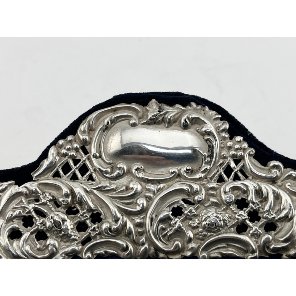A rectangular sterling silver photo frame featuring intricate floral and scroll repousse metalwork mounted on a dark blue velvet easel backing. View VII