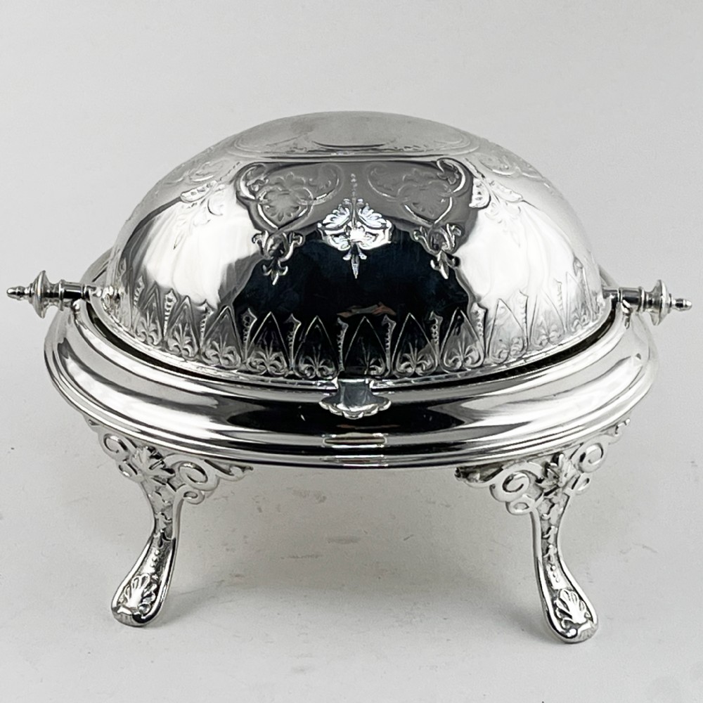 A Victorian silver plated revolving butter dish featuring an engraved domed lid, decorative side pivot handles, and four scrolled legs.