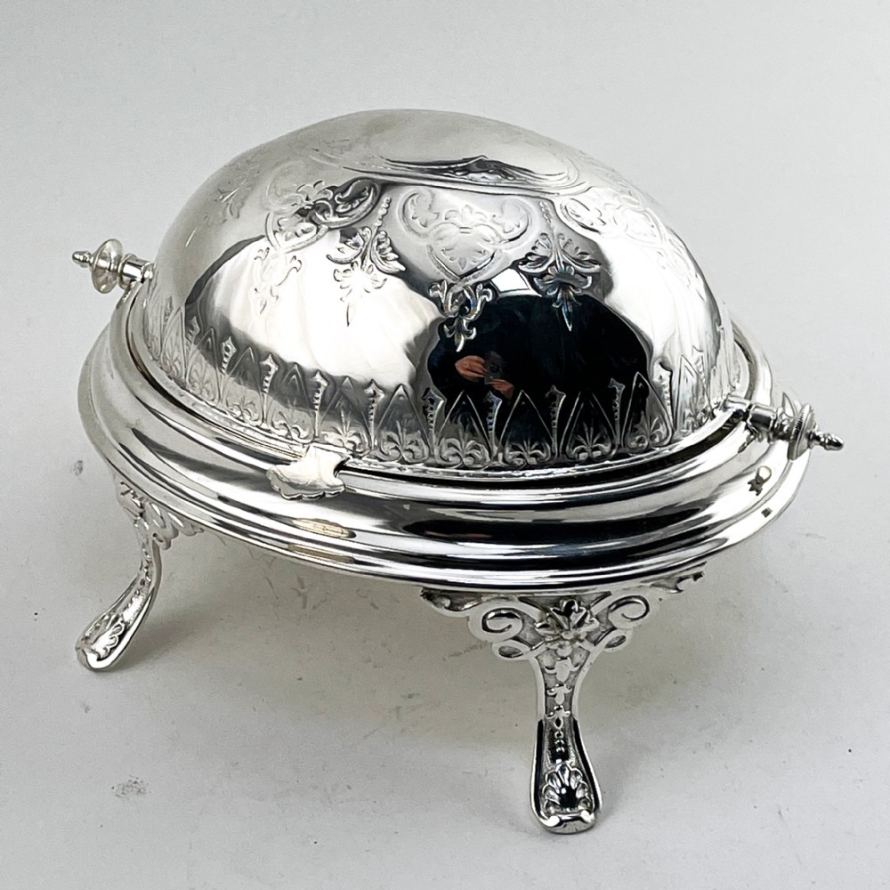 A Victorian silver plated revolving butter dish featuring an engraved domed lid, decorative side pivot handles, and four scrolled legs. View II