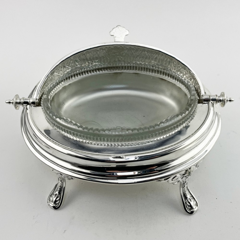 A Victorian silver plated revolving butter dish featuring an engraved domed lid, decorative side pivot handles, and four scrolled legs. View III