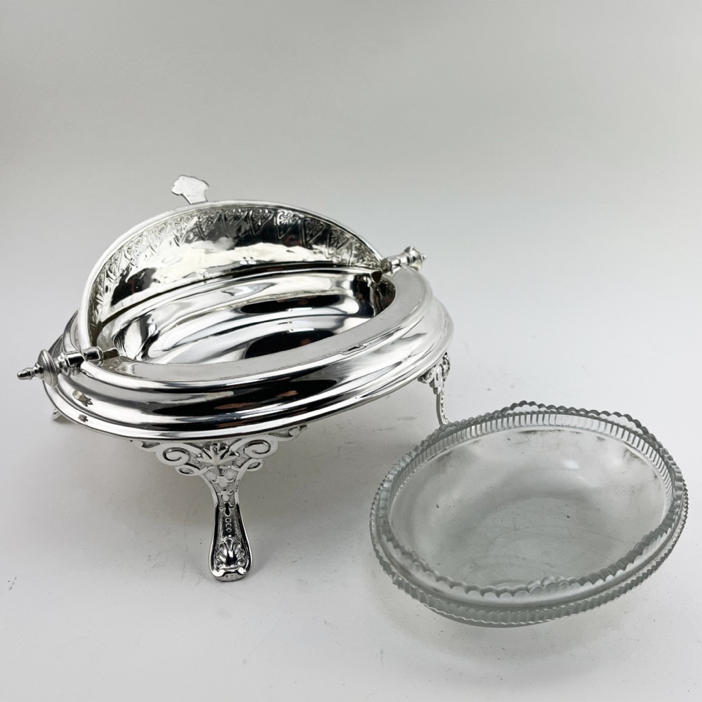 A Victorian silver plated revolving butter dish featuring an engraved domed lid, decorative side pivot handles, and four scrolled legs. View V