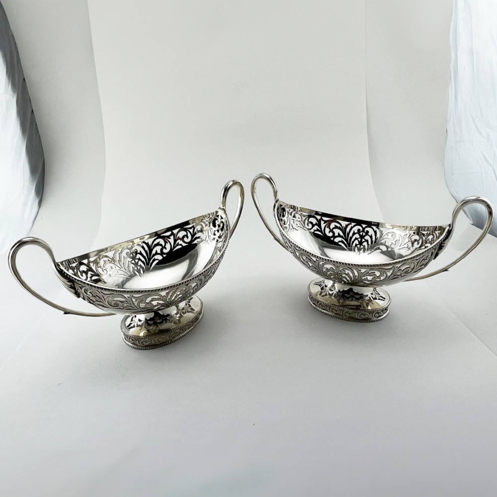 A pair of silver-plated boat-shaped dishes featuring pierced scrollwork borders, high reeded side handles, and oval pedestal bases.