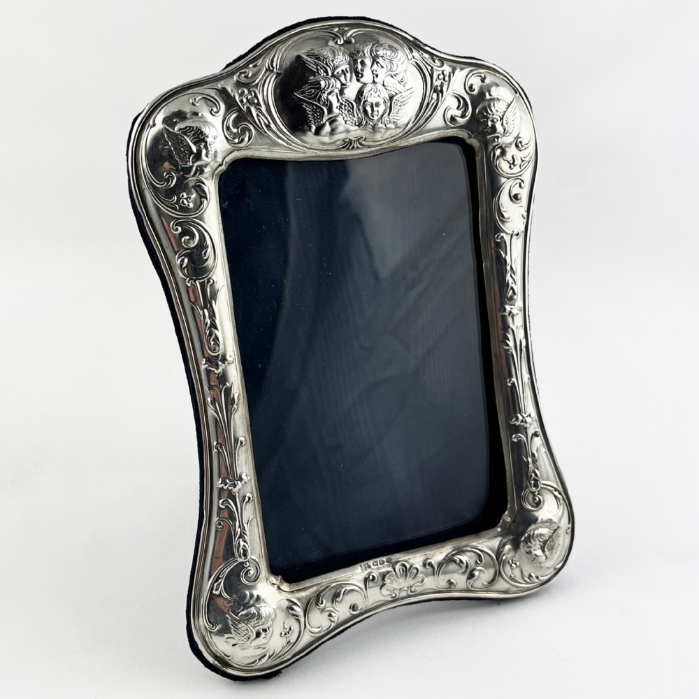 A shaped Victorian sterling silver photograph frame featuring embossed cherub motifs inspired by Reynolds Angels, scrollwork borders, and a dark velvet easel back.