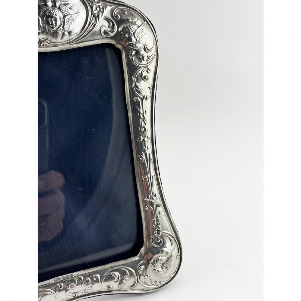 A shaped Victorian sterling silver photograph frame featuring embossed cherub motifs inspired by Reynolds Angels, scrollwork borders, and a dark velvet easel back. View II