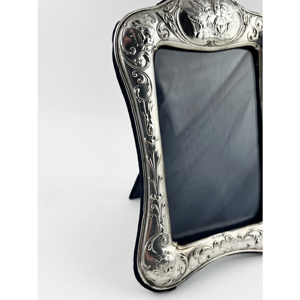 A shaped Victorian sterling silver photograph frame featuring embossed cherub motifs inspired by Reynolds Angels, scrollwork borders, and a dark velvet easel back. View III