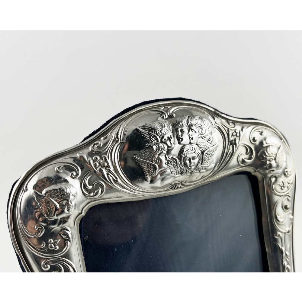 A shaped Victorian sterling silver photograph frame featuring embossed cherub motifs inspired by Reynolds Angels, scrollwork borders, and a dark velvet easel back. View IV