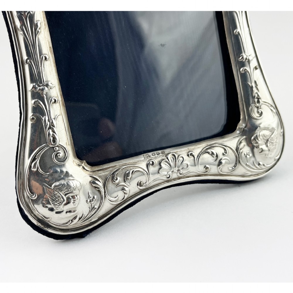 A shaped Victorian sterling silver photograph frame featuring embossed cherub motifs inspired by Reynolds Angels, scrollwork borders, and a dark velvet easel back. View V