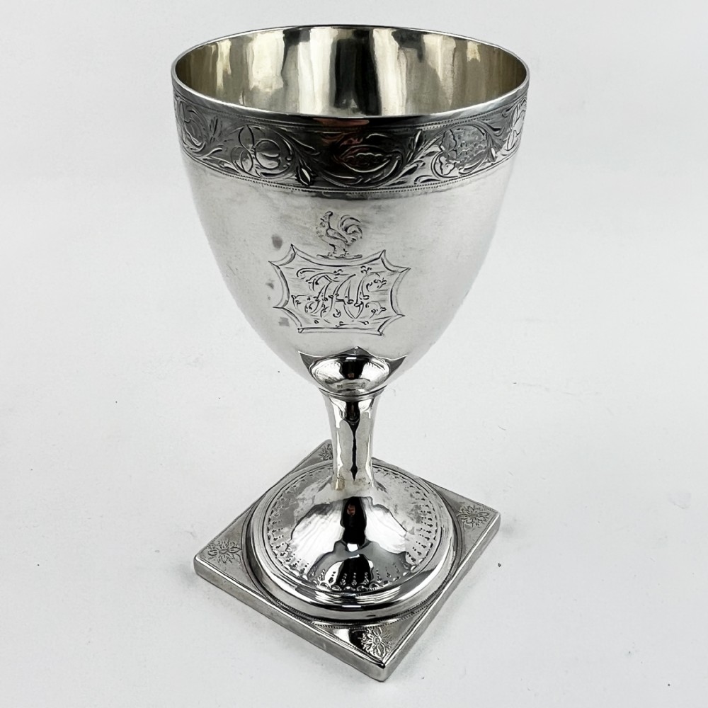 A Georgian sterling silver goblet featuring a vase-shaped bowl with engraved crest and foliate border set upon a square plinth base.
