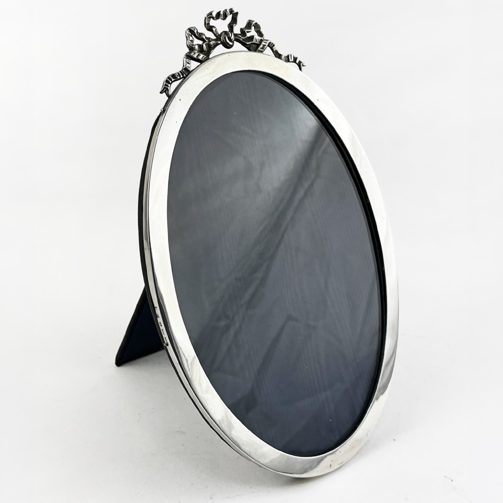 An oval sterling silver photo frame with a decorative ribbon crest at the top and an easel back stand.