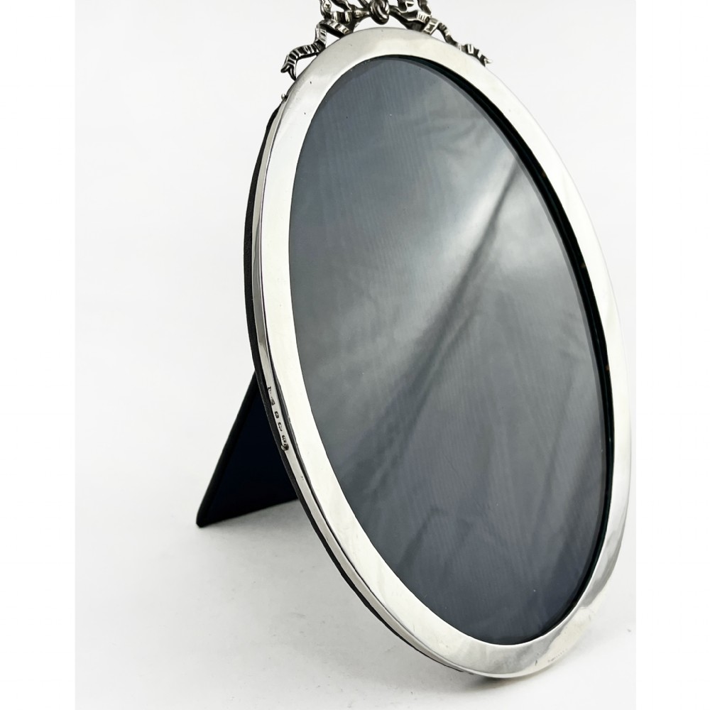 An oval sterling silver photo frame with a decorative ribbon crest at the top and an easel back stand. View III