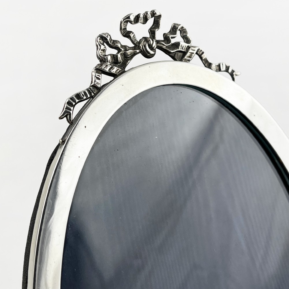 An oval sterling silver photo frame with a decorative ribbon crest at the top and an easel back stand. View IV