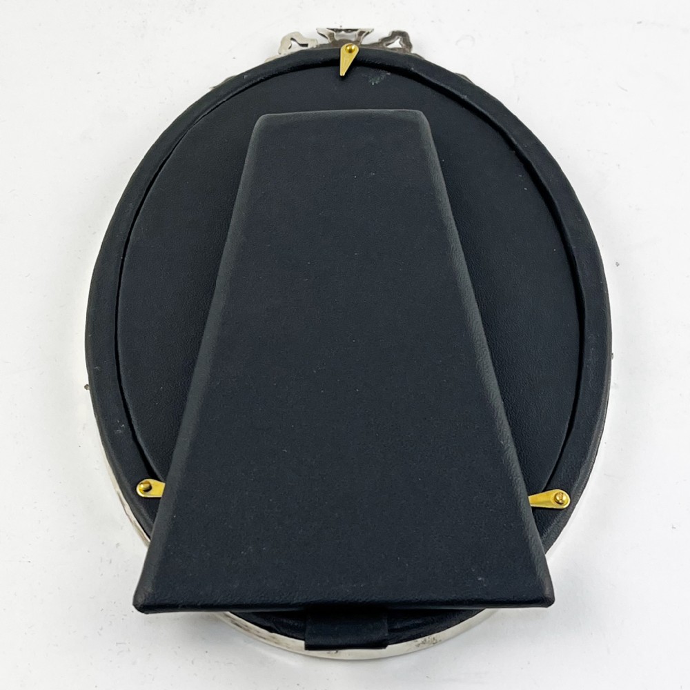 An oval sterling silver photo frame with a decorative ribbon crest at the top and an easel back stand. View VI