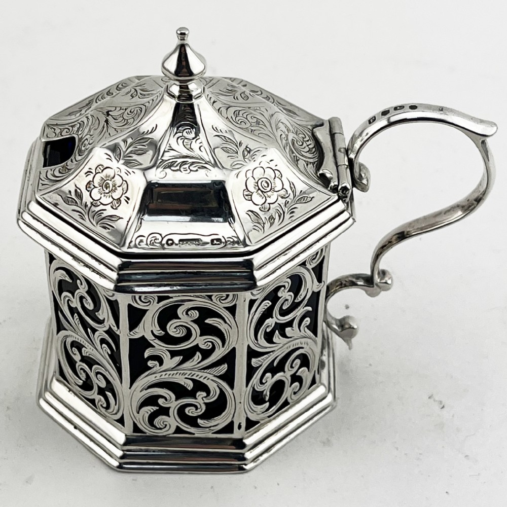 An octagonal Victorian sterling silver mustard pot featuring pierced foliate scrollwork, an engraved hinged lid with finial, a scroll handle, and an inner cobalt blue glass liner.