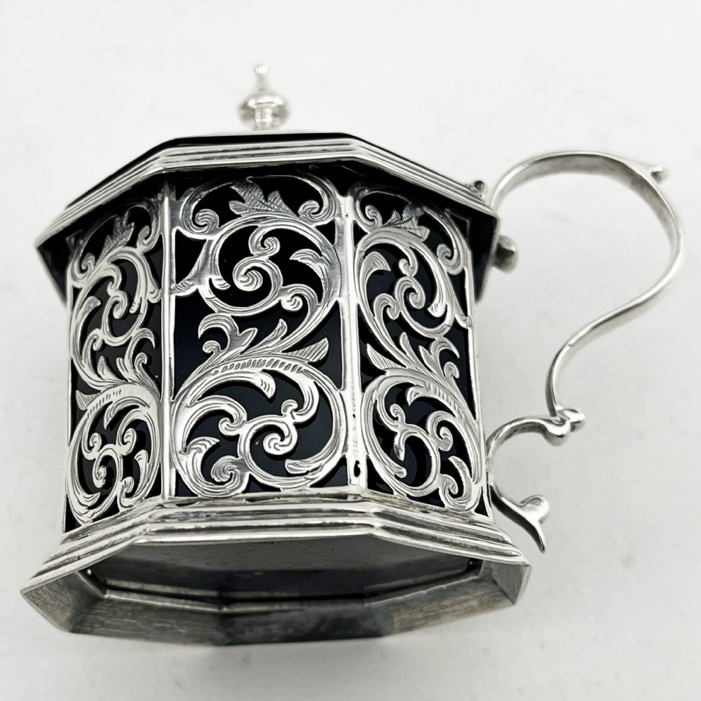An octagonal Victorian sterling silver mustard pot featuring pierced foliate scrollwork, an engraved hinged lid with finial, a scroll handle, and an inner cobalt blue glass liner. View V