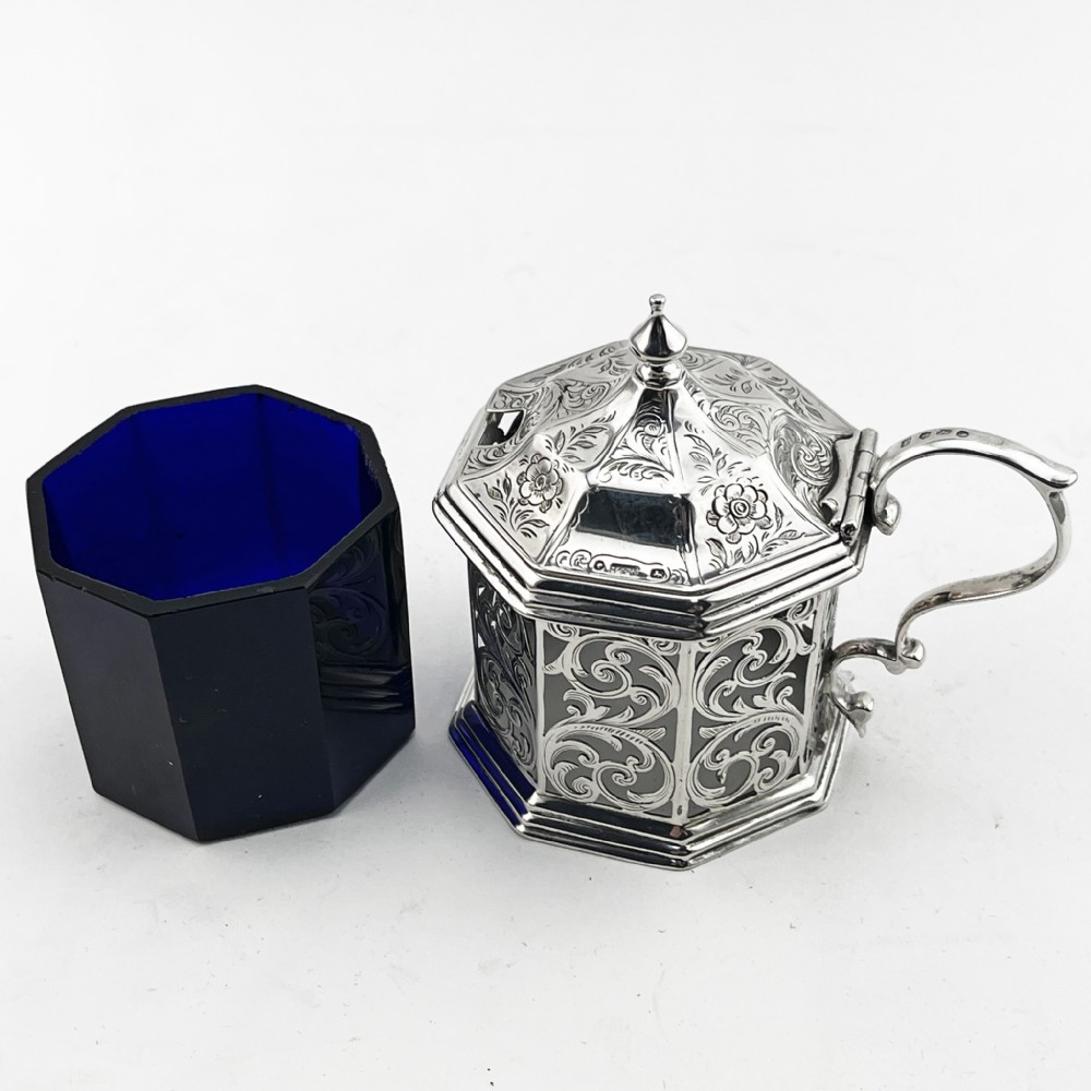 An octagonal Victorian sterling silver mustard pot featuring pierced foliate scrollwork, an engraved hinged lid with finial, a scroll handle, and an inner cobalt blue glass liner. View VII