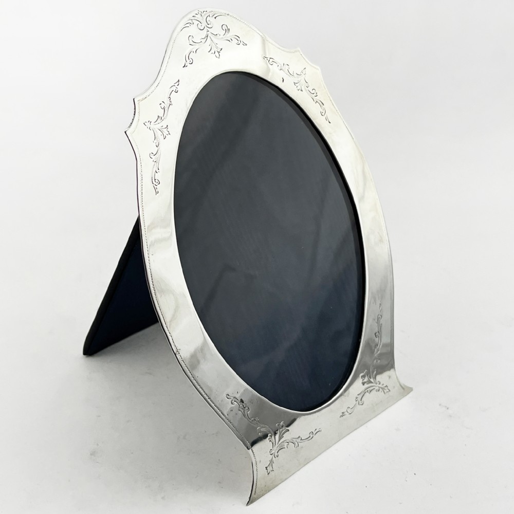 A sterling silver photo frame with an oval opening, engraved foliate detailing, a shaped crest, and a dark velvet easel backing.