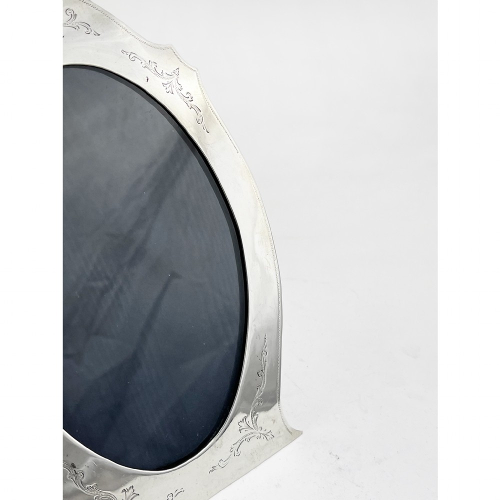 A sterling silver photo frame with an oval opening, engraved foliate detailing, a shaped crest, and a dark velvet easel backing. View II