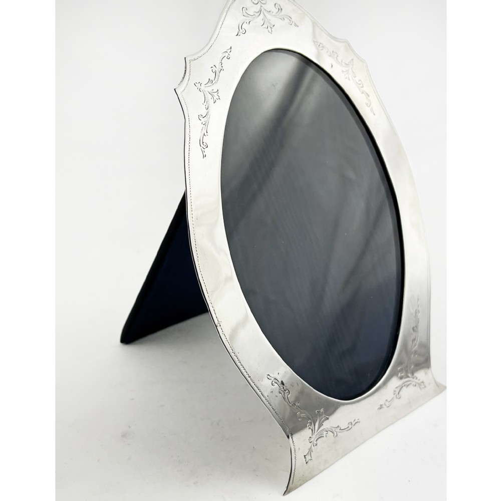 A sterling silver photo frame with an oval opening, engraved foliate detailing, a shaped crest, and a dark velvet easel backing. View III