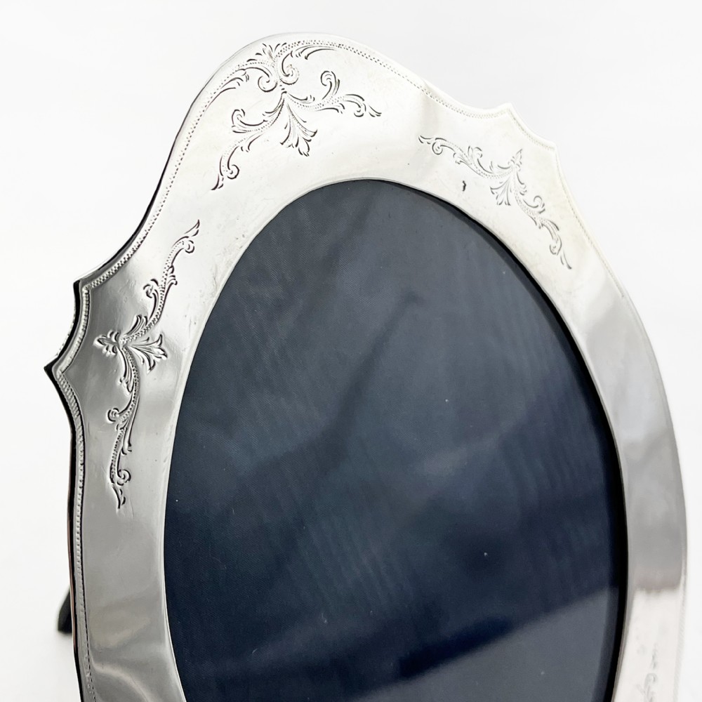 A sterling silver photo frame with an oval opening, engraved foliate detailing, a shaped crest, and a dark velvet easel backing. View IV