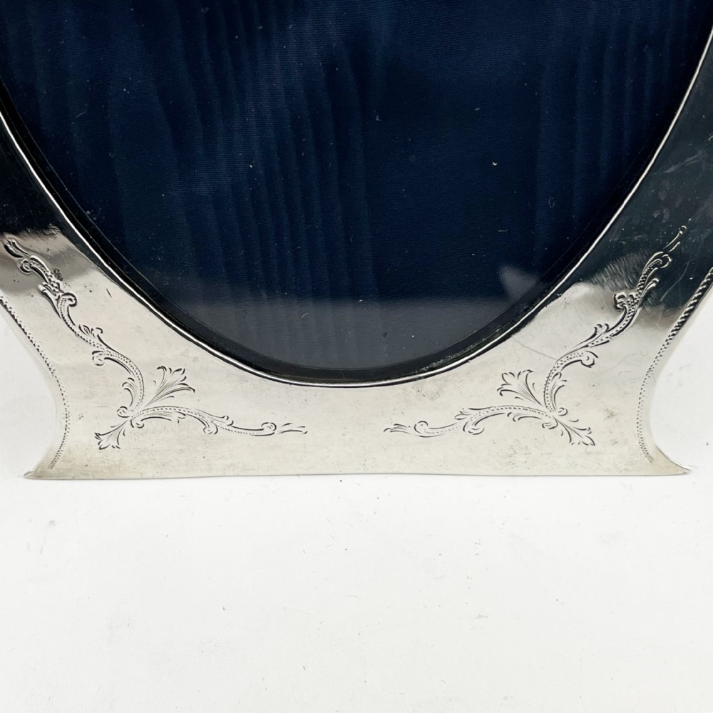 A sterling silver photo frame with an oval opening, engraved foliate detailing, a shaped crest, and a dark velvet easel backing. View V