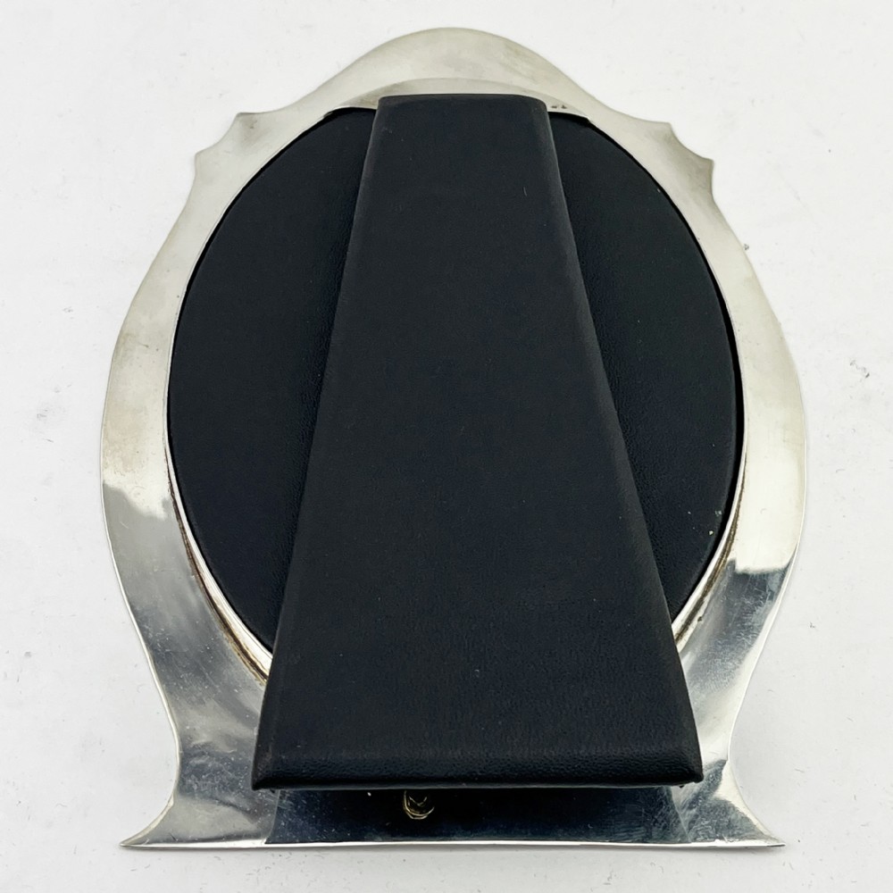 A sterling silver photo frame with an oval opening, engraved foliate detailing, a shaped crest, and a dark velvet easel backing. View VII