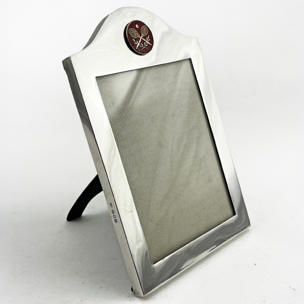 A sterling silver portrait photo frame featuring an arched top decorated with a round red enamel crest depicting crossed tennis racquets and a ball, supported by a rear easel stand.