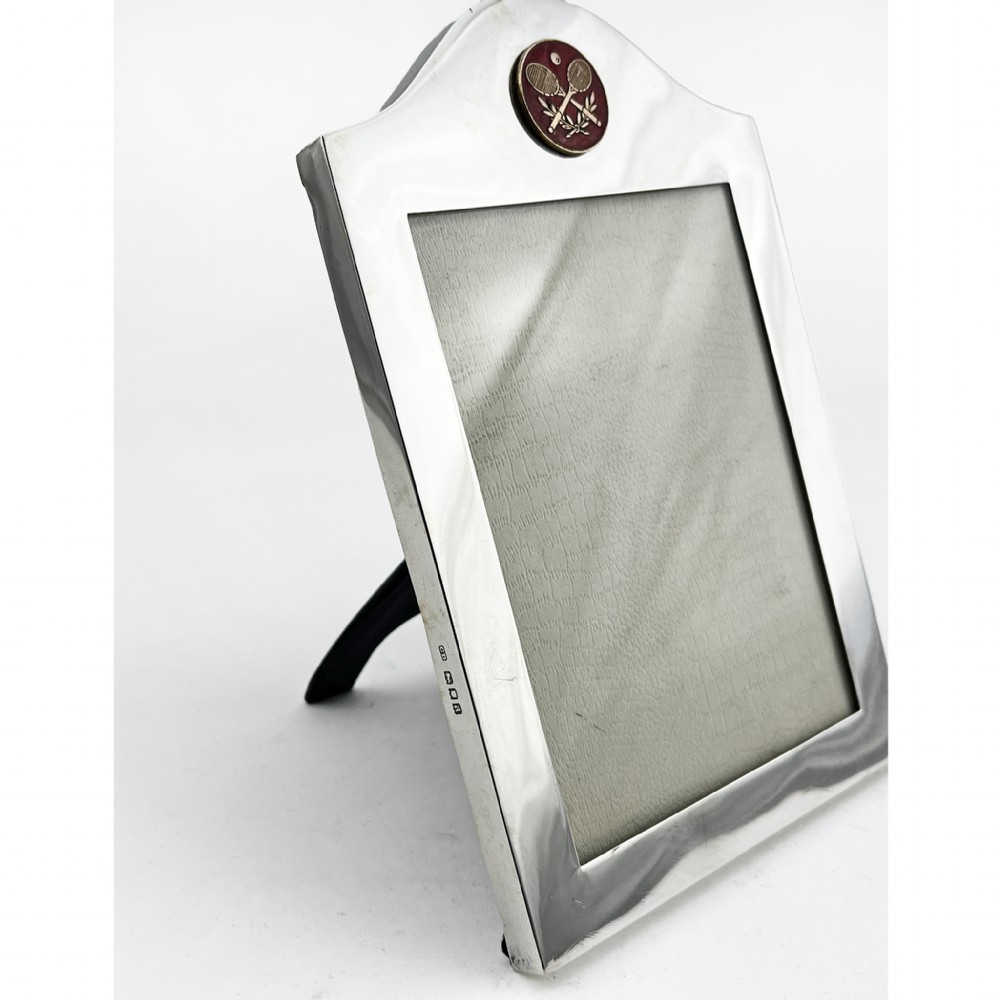 A sterling silver portrait photo frame featuring an arched top decorated with a round red enamel crest depicting crossed tennis racquets and a ball, supported by a rear easel stand. View IV