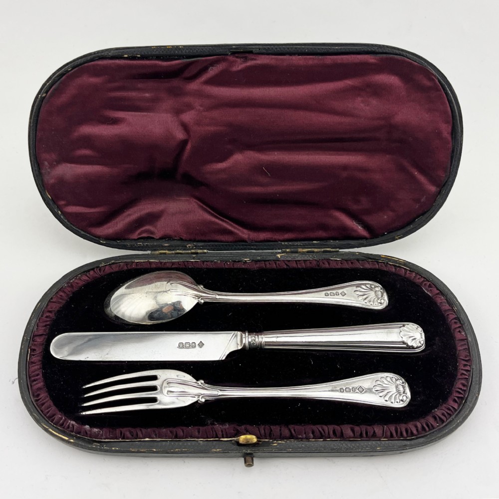 A three-piece Victorian sterling silver christening set featuring a spoon, knife, and fork arranged inside an open black leatherette presentation box lined with plum silk and velvet.