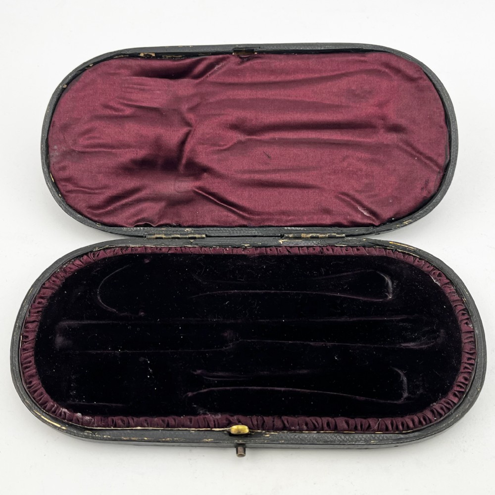 A three-piece Victorian sterling silver christening set featuring a spoon, knife, and fork arranged inside an open black leatherette presentation box lined with plum silk and velvet. View V
