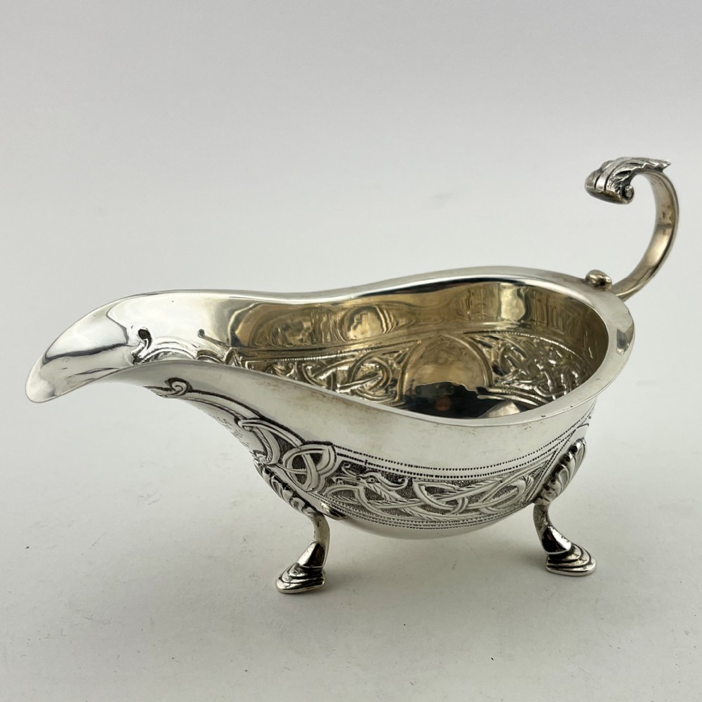 An Irish sterling silver sauce boat decorated with chased Celtic knotwork, featuring a scroll handle and three hoof feet.