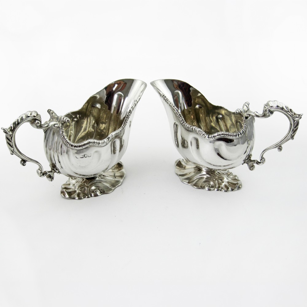 A pair of polished sterling silver sauce boats featuring fluted bodies, scroll handles, and decorated pedestal bases.