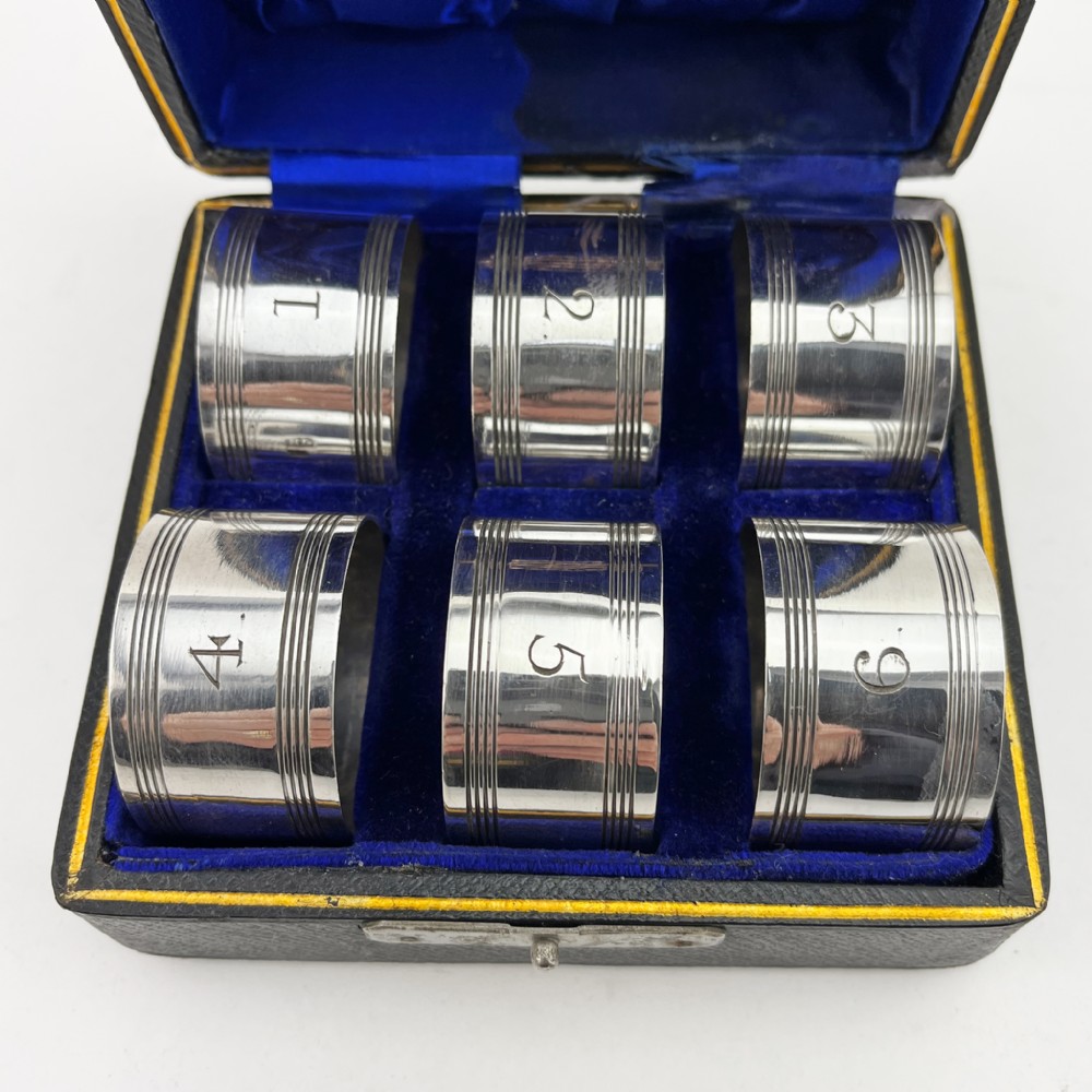 A set of six silver-plated napkin rings numbered one to six with reeded borders, stored in an open black case lined with blue silk and velvet. View II