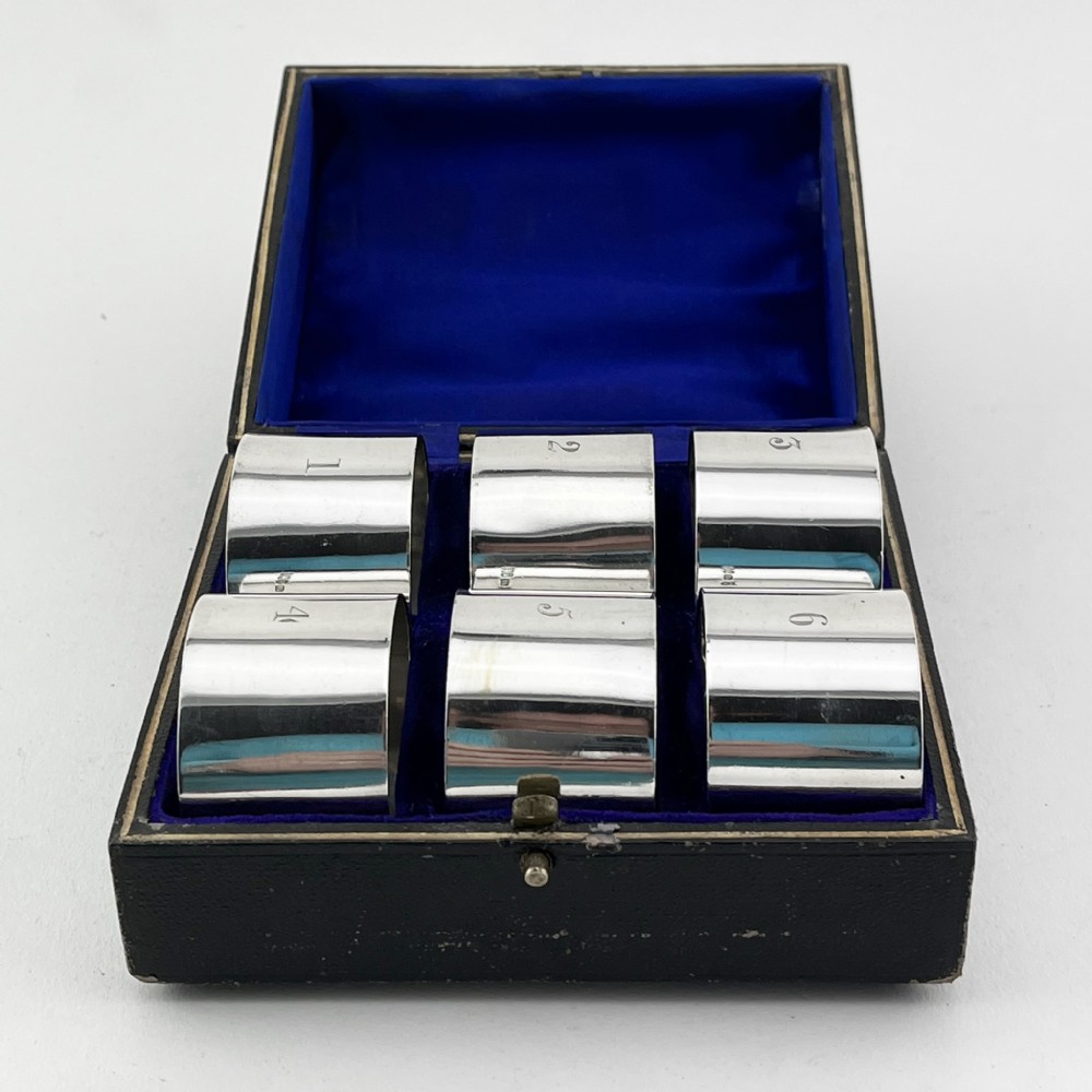 A black fitted presentation box open to display six plain sterling silver napkin rings numbered one through six on cobalt blue velvet lining.