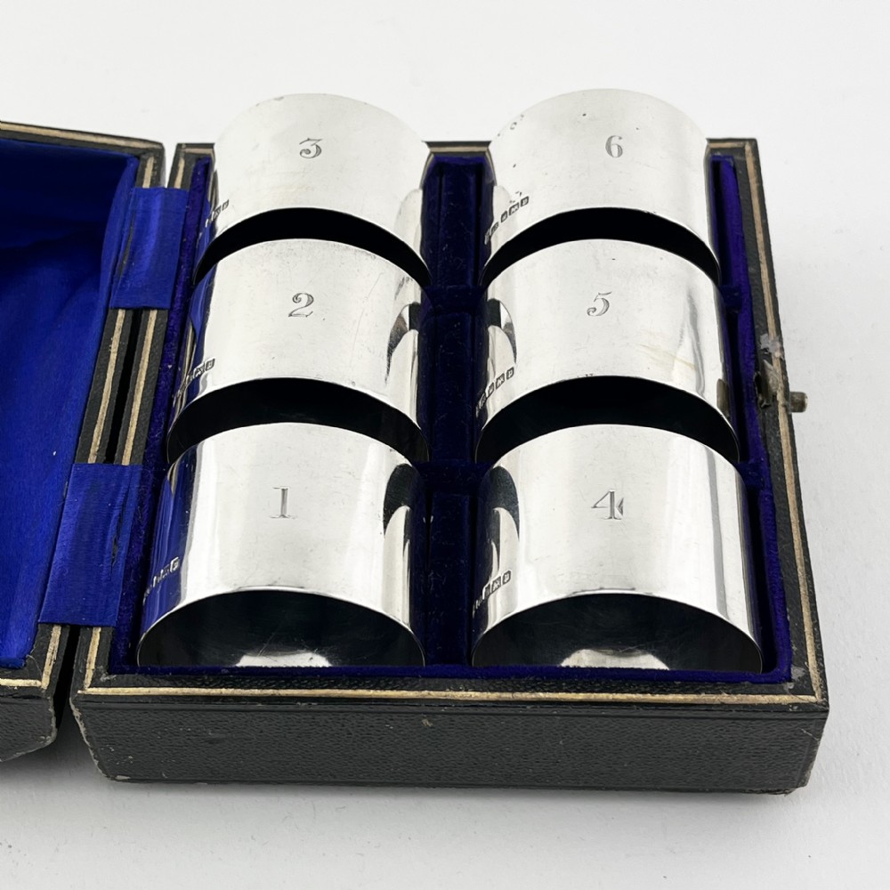 A black fitted presentation box open to display six plain sterling silver napkin rings numbered one through six on cobalt blue velvet lining. View II