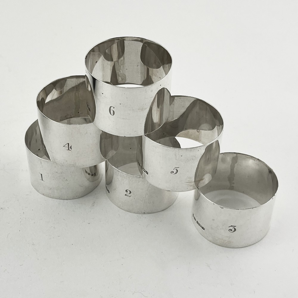 A black fitted presentation box open to display six plain sterling silver napkin rings numbered one through six on cobalt blue velvet lining. View III