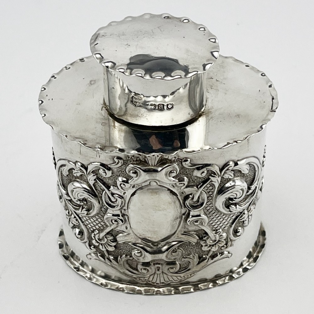 An oval Victorian sterling silver tea caddy featuring chased scrollwork decoration, a blank central cartouche, a scalloped border, and a pull-off cap lid.