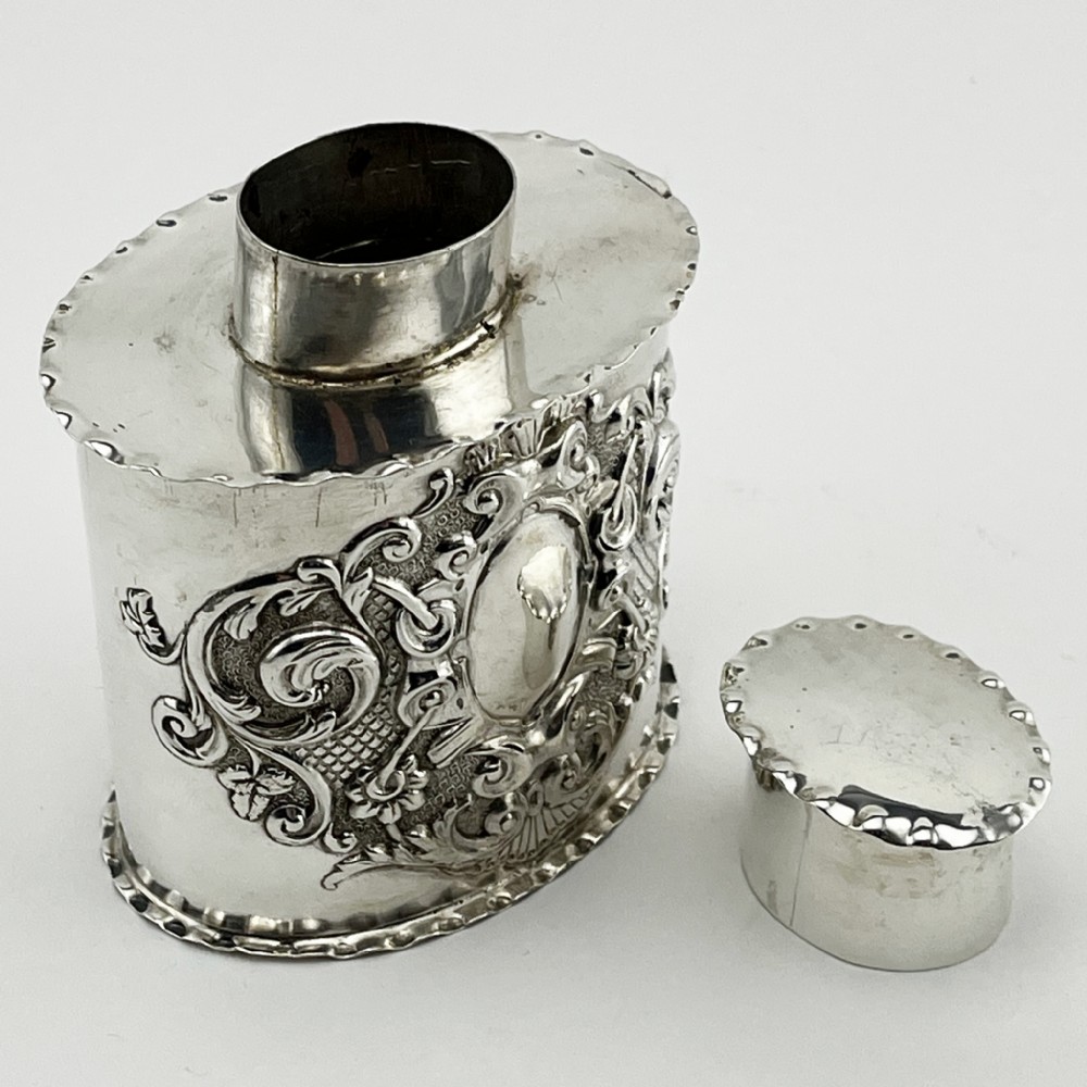 An oval Victorian sterling silver tea caddy featuring chased scrollwork decoration, a blank central cartouche, a scalloped border, and a pull-off cap lid. View IV