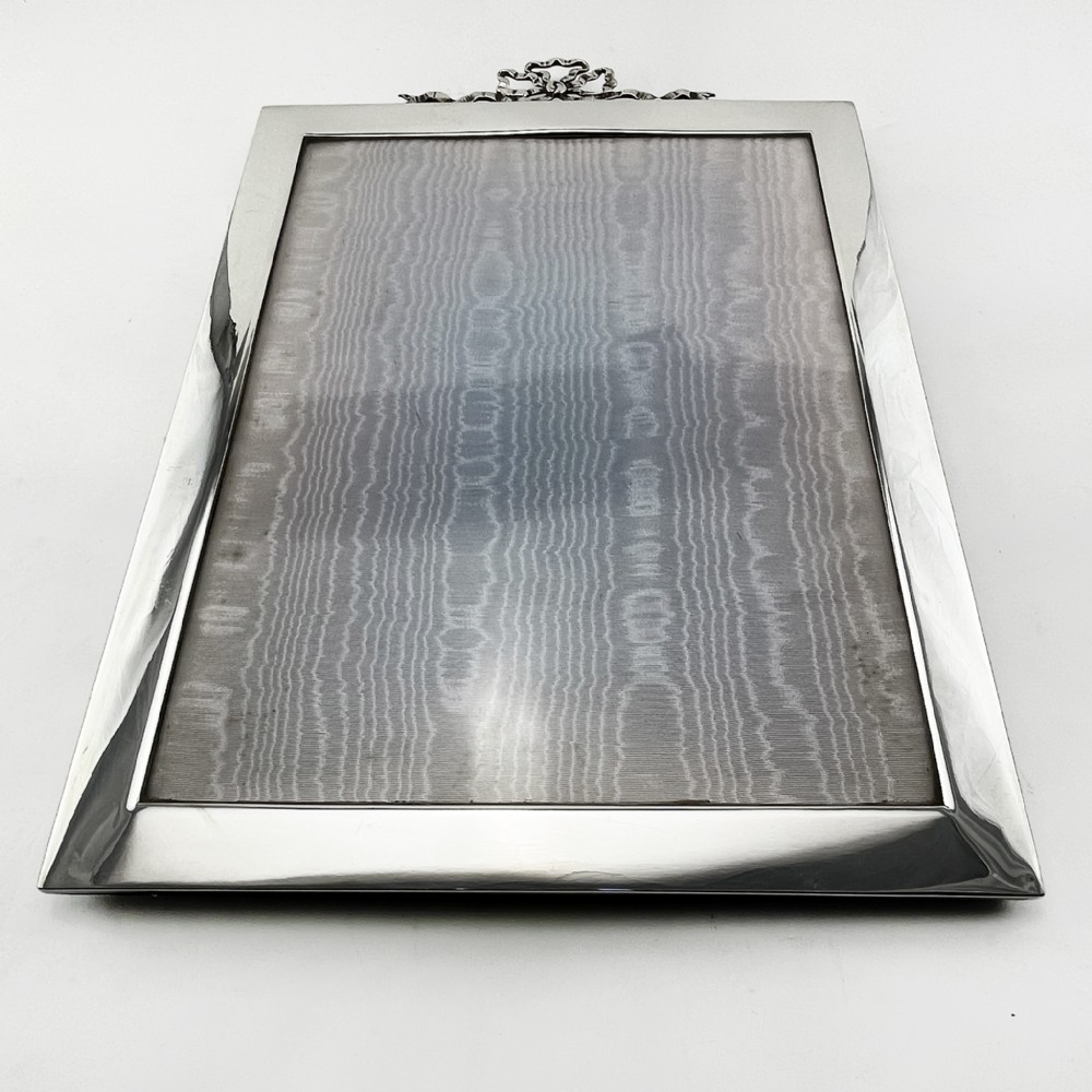 A rectangular sterling silver photo frame featuring a plain polished border topped with a bow ribbon motif. View V