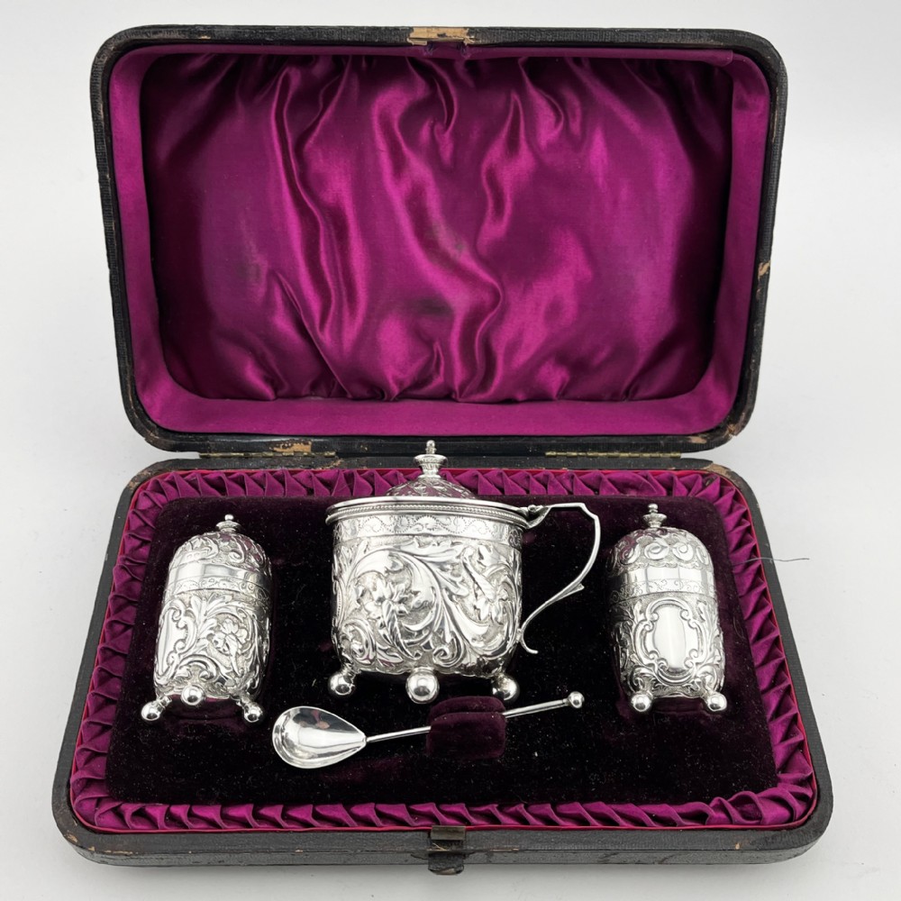 A Victorian sterling silver three-piece condiment set with floral repousse engraving housed inside an open velvet-lined presentation case.
