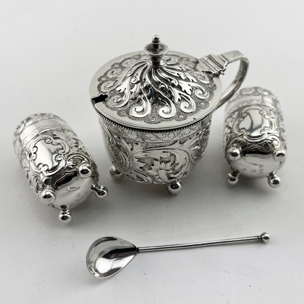 A Victorian sterling silver three-piece condiment set with floral repousse engraving housed inside an open velvet-lined presentation case. View II