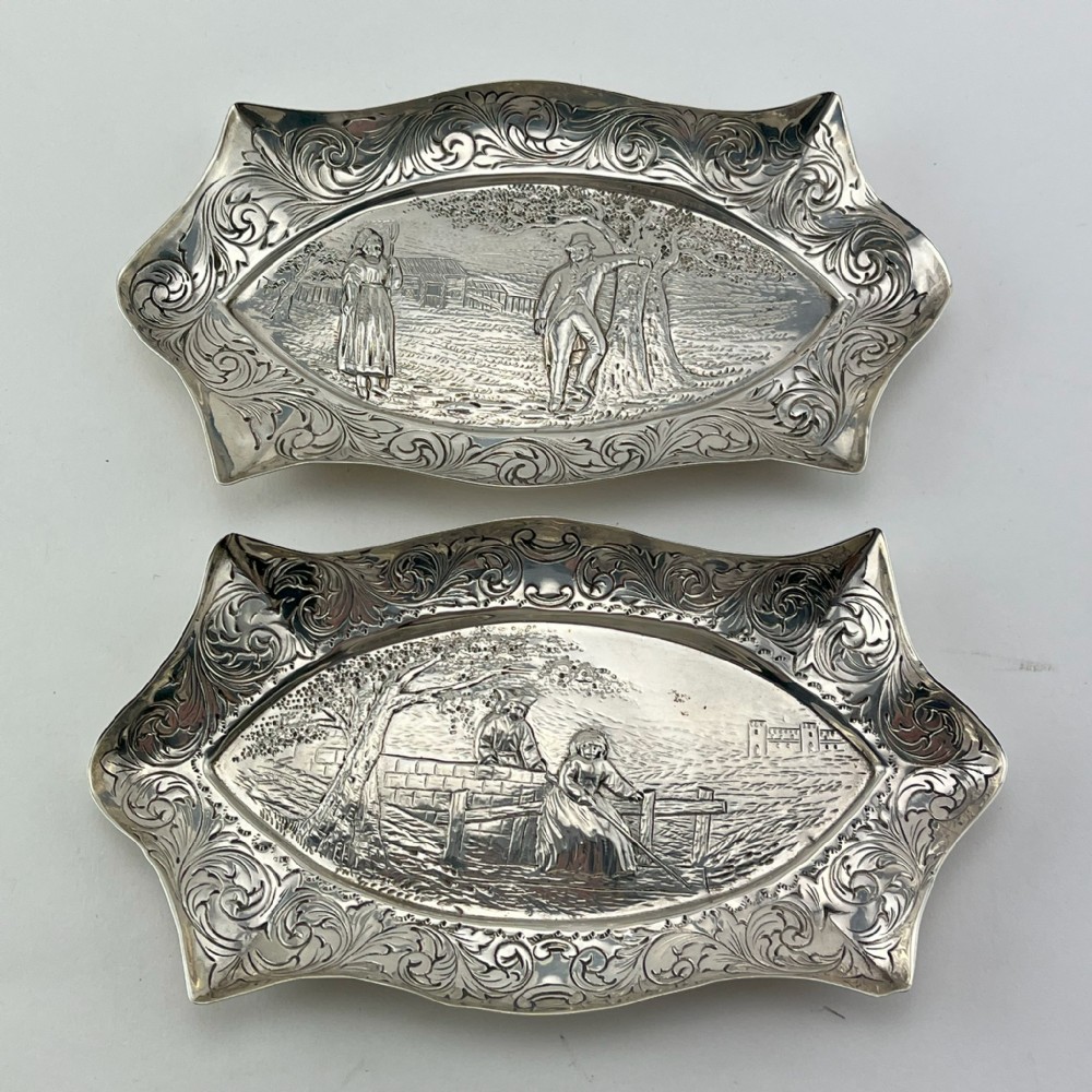 A pair of Victorian sterling silver dishes featuring relief-embossed pastoral scenes with ornate scrolled borders.
