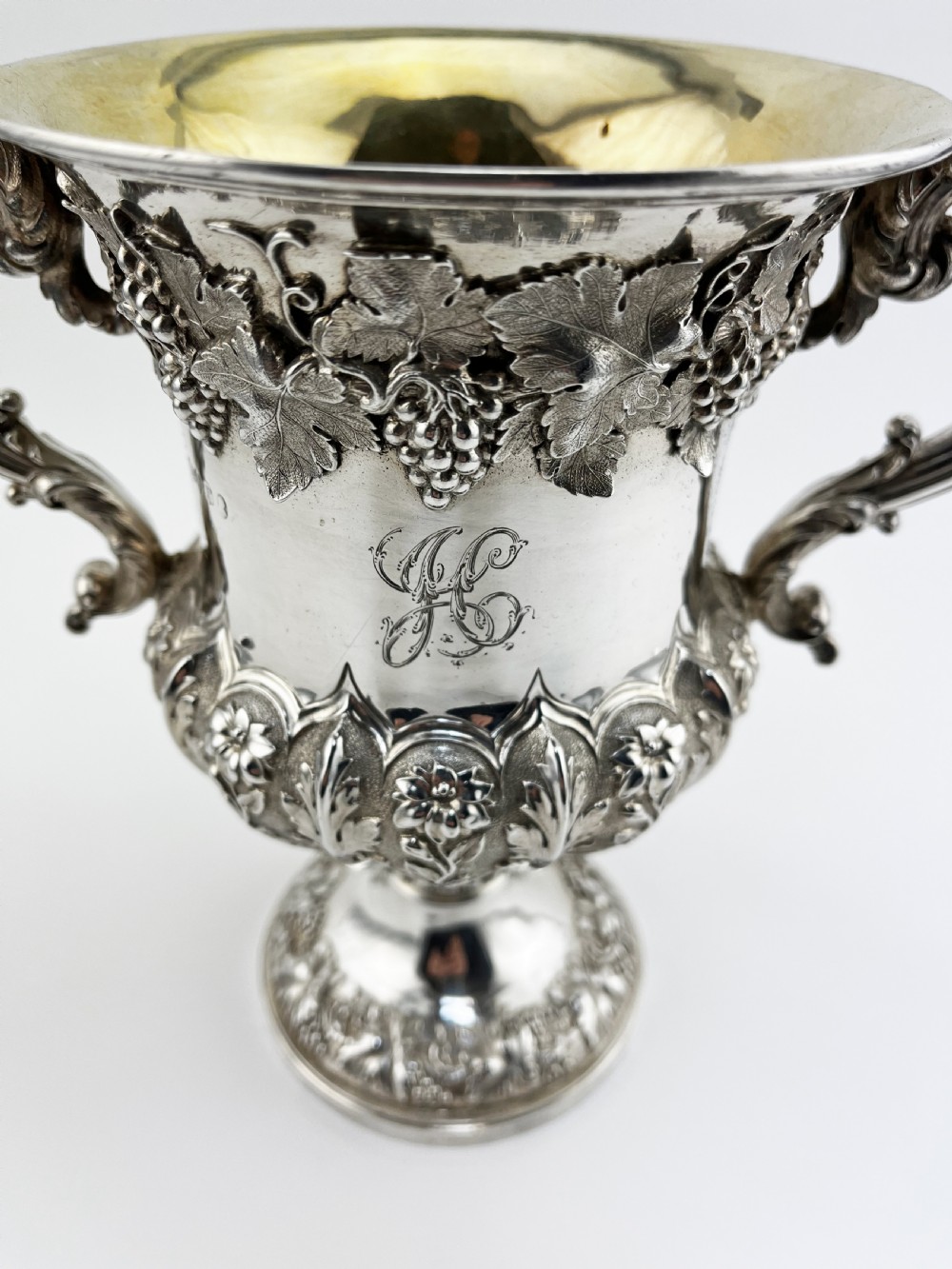 A William IV sterling silver trophy cup featuring raised grapevine repousse motifs, double scroll handles, and a gilt interior. View IV