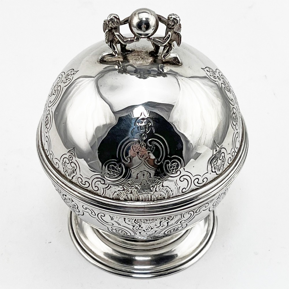 A spherical George III sterling silver soap box featuring engraved scrollwork and a finial shaped like two cherubs holding an orb.
