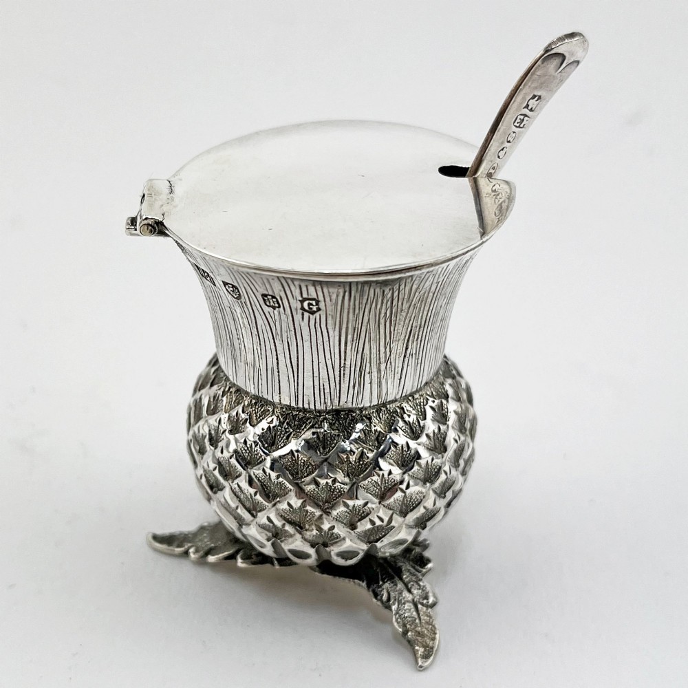 A sterling silver mustard pot styled in the shape of a Scottish thistle with a flat hinged lid and an inserted matching silver spoon.