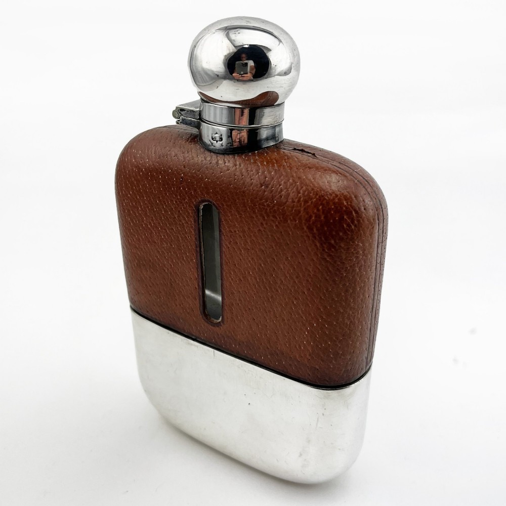 A silver plated hip flask featuring a textured brown leather upper section with a vertical sight window and a polished domed cap.