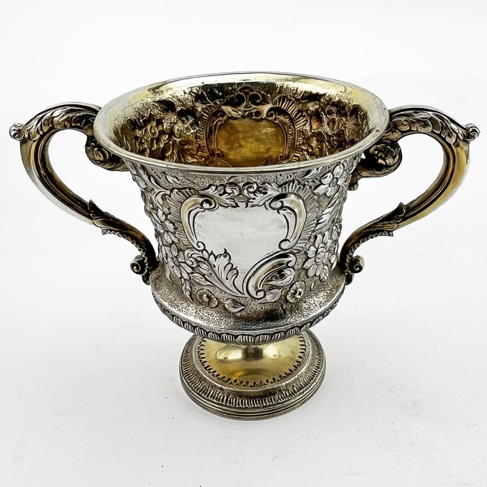 A George III sterling silver campana-shaped cup featuring chased floral motifs, a vacant cartouche, twin scroll handles, and a gilt interior.
