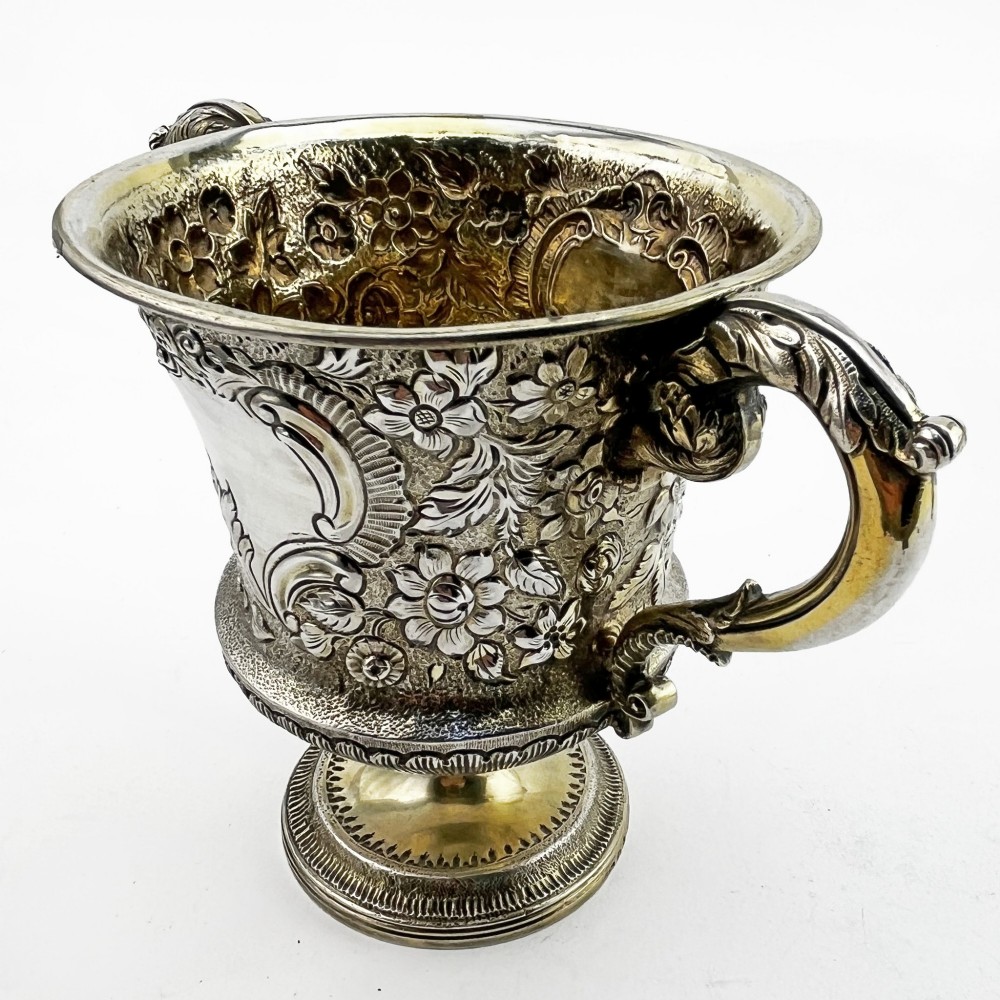 A George III sterling silver campana-shaped cup featuring chased floral motifs, a vacant cartouche, twin scroll handles, and a gilt interior. View II