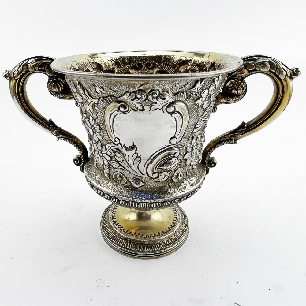 A George III sterling silver campana-shaped cup featuring chased floral motifs, a vacant cartouche, twin scroll handles, and a gilt interior. View III